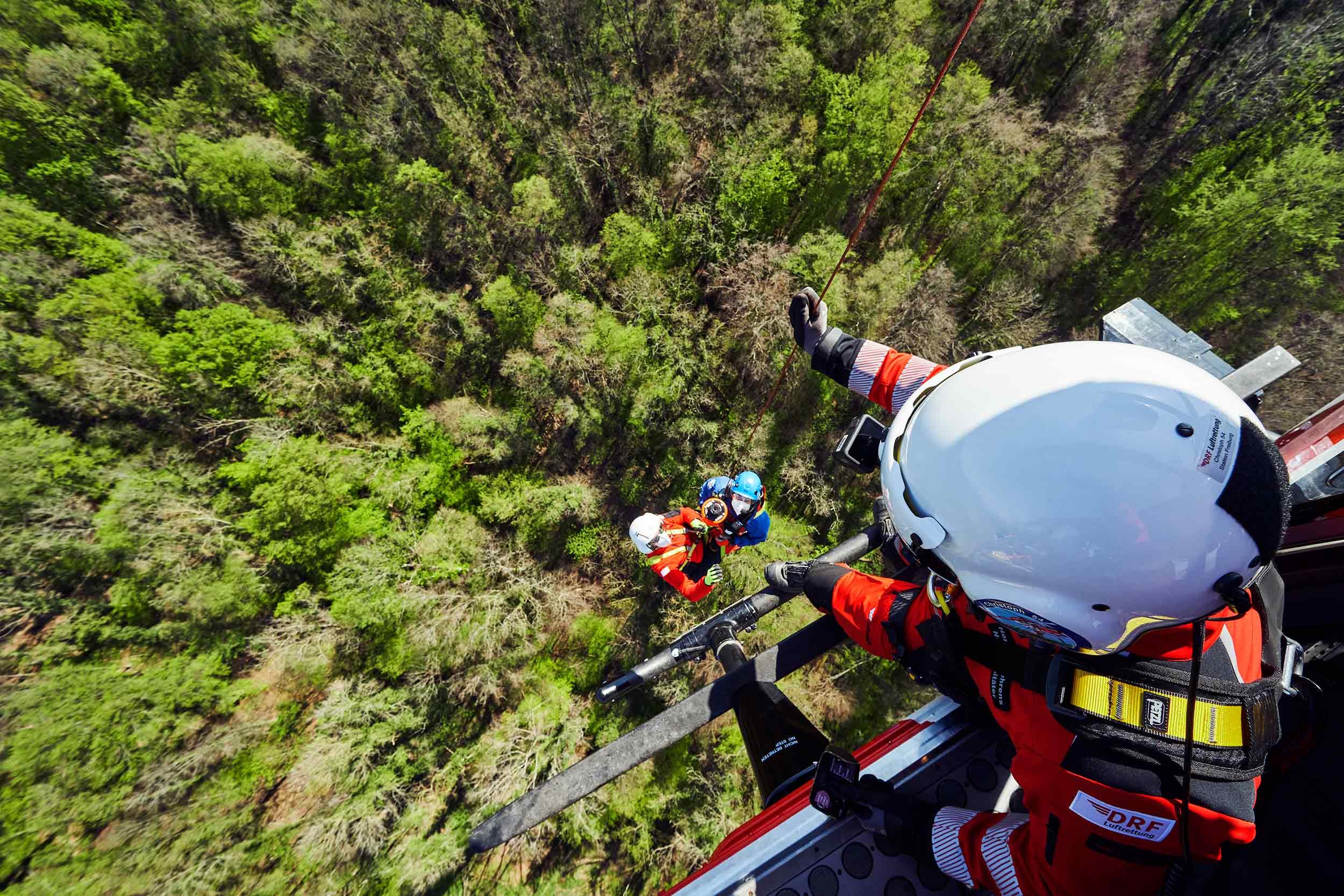 View from the open side door of the helicopter of two people being winched down with the rescue winch.
